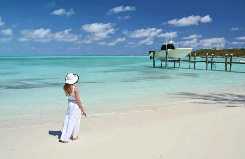 Frau in weißem Kleid und Sonnenhut spaziert an einem weißen Sandstrand auf den Bahamas.