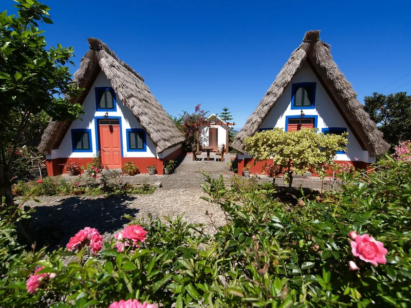 Traditionelle Santana-Häuser mit Reetdach und bunten Fassaden in einem blühenden Garten auf Madeira, Portugal.