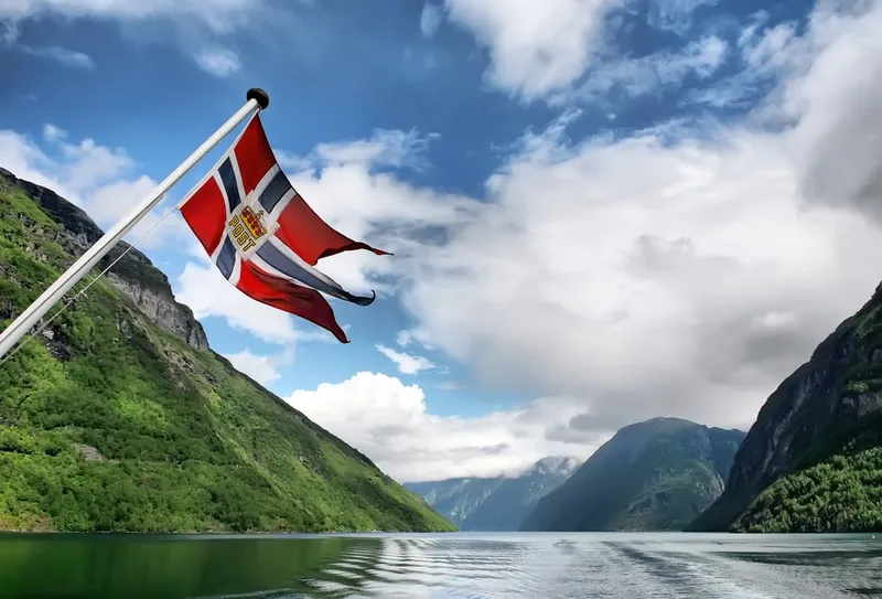 Norwegische Flagge weht im Wind auf einem Schiff inmitten eines grünen norwegischen Fjords.