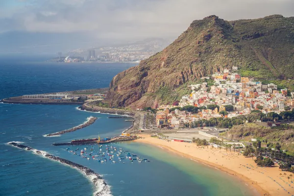 Küstenansicht von Teneriffa mit goldenem Sandstrand, buntem Bergdorf und Blick auf Santa Cruz im Hintergrund.